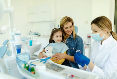 Happy mother and child seeing pediatric dentist. Oral hygienist with jaw model teaching patients to brush teeth properly to prevent caries, keep teeth clean and healthy and have good beautiful smile.